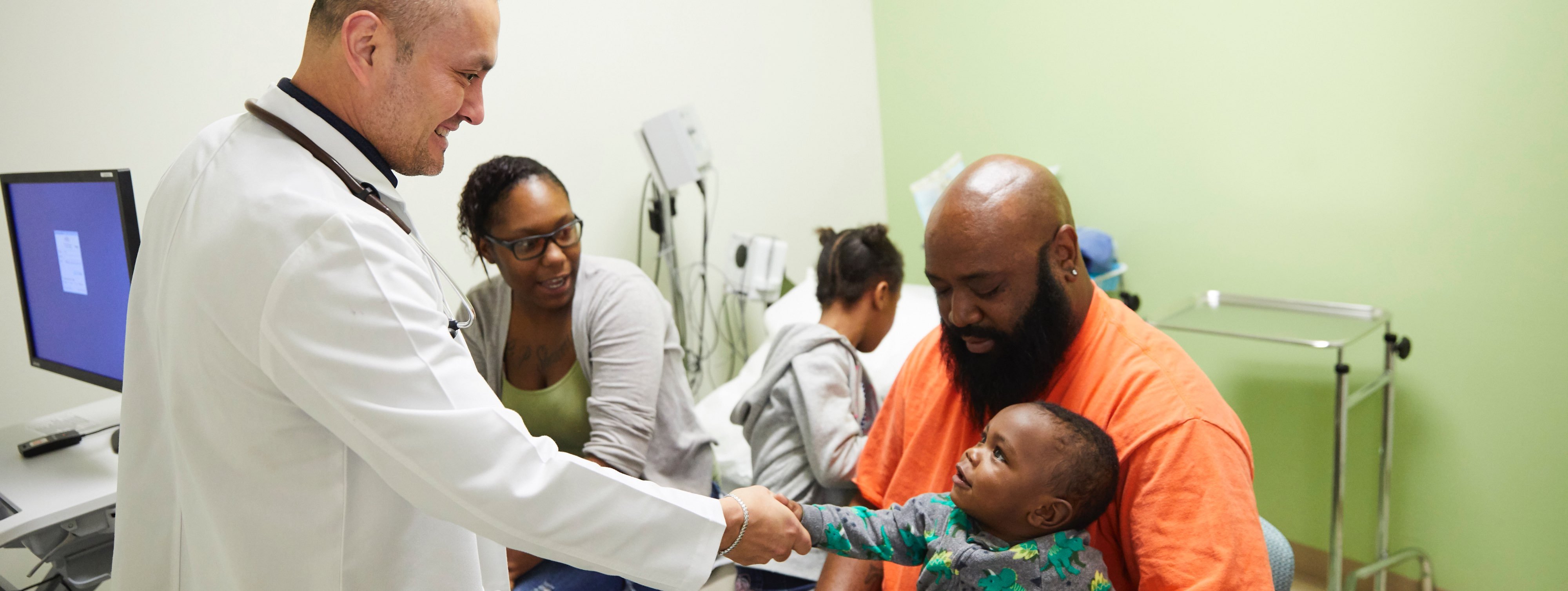 Médico latino sosteniendo la mano de un bebé de color que se encuentra sentado en el regazo de su padre mientras su madre los mira